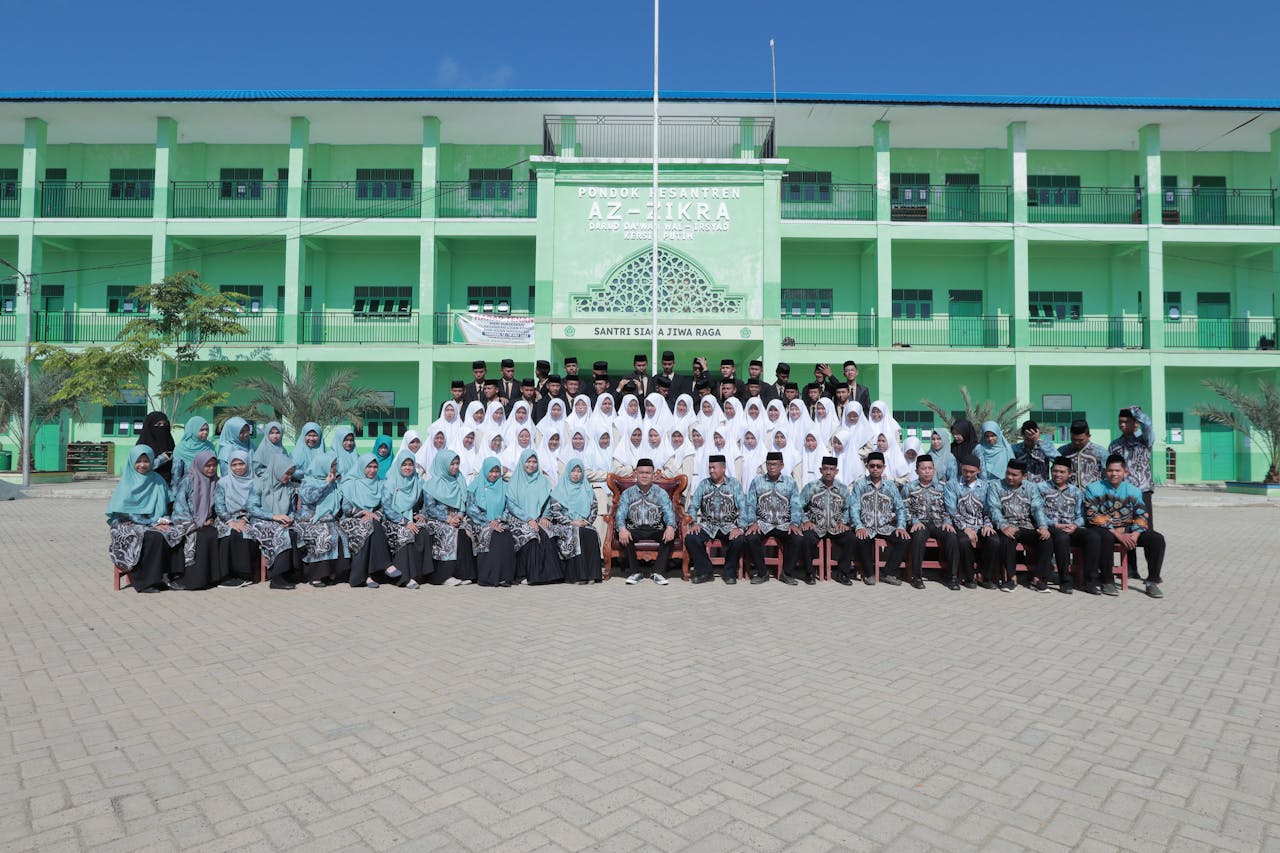 Students and teachers posing for a graduation photo at an Islamic school courtyard under clear skies.