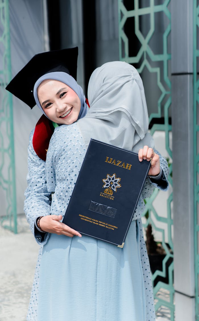 Two women celebrating graduation with a joyful hug, wearing hijabs and holding diploma.