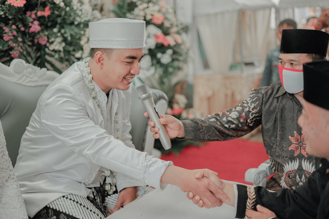 Groom in traditional attire shaking hands during wedding ceremony with floral decor.
