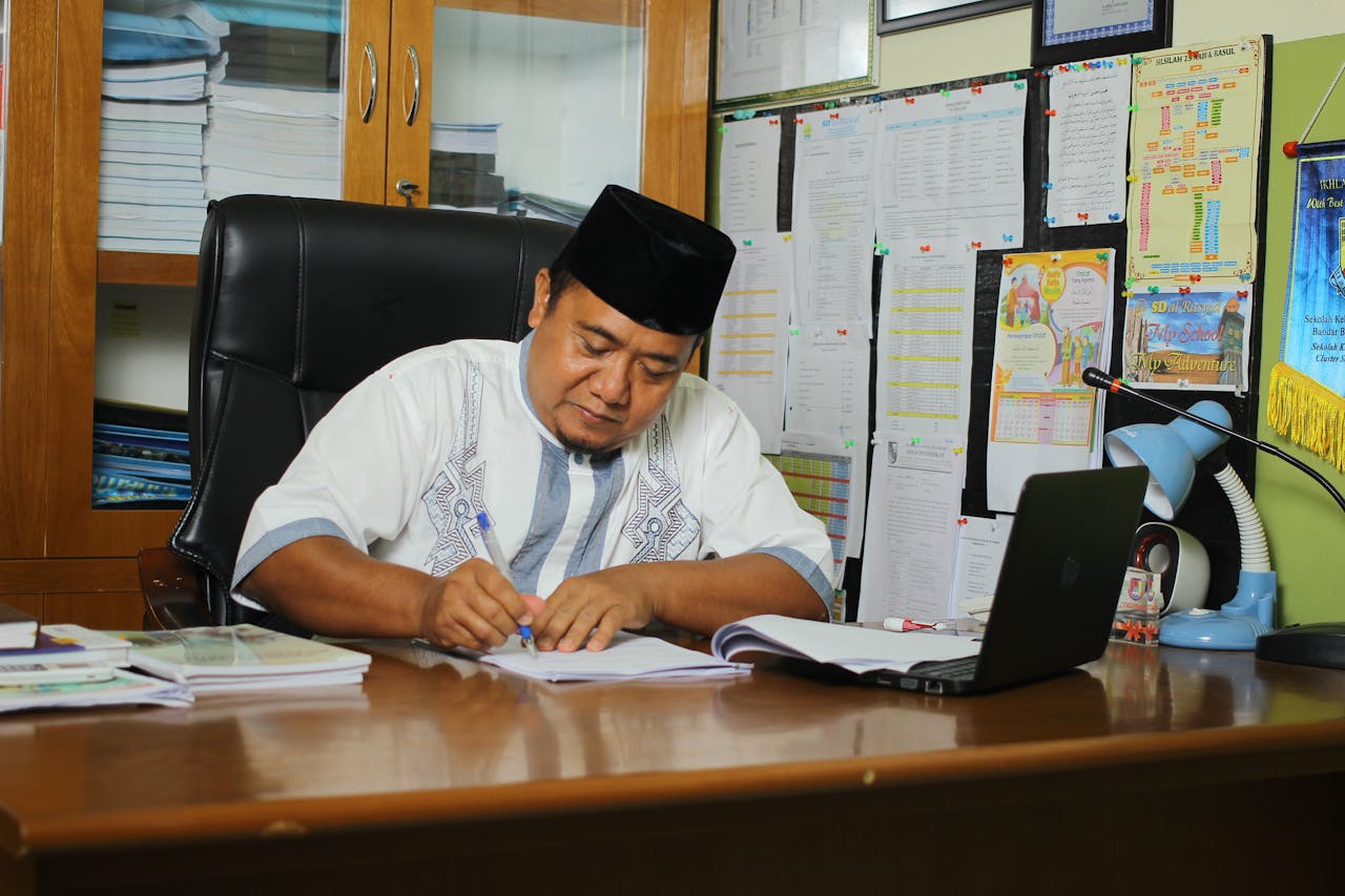 Indonesian educator writing at his desk, focused on paperwork in an office setting.